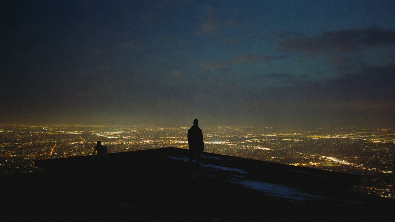 A lone figure on a high overlook, watching a city glow with warm light at dusk